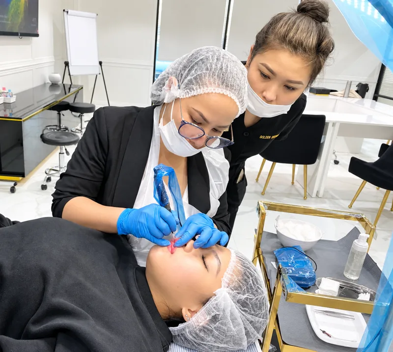 A student tattooing brows on a real model, Tina beside her